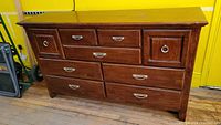Front view of wooden dresser with drawer configuration visible. Medium-brown wood with metal ring and cup handles. Some surface scratches on top.