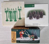 Photo showing three boxes: one contains green street lamps, one contains four twinkling lit green shrubs, and one with Seasons Whimsy figurines featuring a reindeer and gift sack.