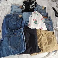Top view of multiple pairs of pants and shorts laid out, showing various colors and styles including denim, khaki, black, and printed fabric shorts