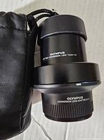 Olympus 1.7x Tele Conversion Lens TCON-17C with lens adapter CLA-1 and black drawstring carry bag next to them.