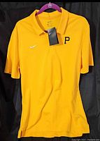 Front view of the yellow Nike Dri-Fit polo shirt with Pittsburgh Pirates 'P' logo and Nike swoosh, hanging on a purple hanger against black background.
