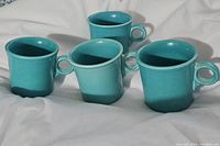Four teal Fiestaware mugs arranged on white fabric, showing handles and overall shape.