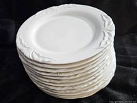 Stack of six white ceramic plates with embossed leaf pattern along edges, viewed from the side showing thickness and relief design.