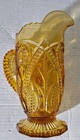 Side view of amber glass pedestal pitcher showing embossed pattern, scalloped rim, and ribbed handle.