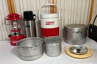 Whole camping collection displayed on table: Coleman lantern, thermos, drink cooler, aluminum cookware pots and lids