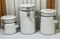 Three white ceramic kitchen storage canisters of varying sizes displayed side by side with attached metal scoops
