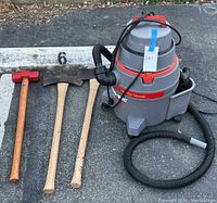 Photo showing wet/dry vacuum with hose and accessories, sledgehammer, and two long handled axes side by side on asphalt.
