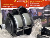 Close up of one black metal landscape light with curved protective cover and frosted diffuser, label showing 11 watt.