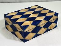 Front angled view of a rectangular wood jewelry box with alternating tan and dark blue diamond-shaped inlays forming a geometric pattern.