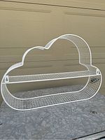White metal cloud-shaped wire mesh shelf shown against a wall and placed on a surface to show shape and size.