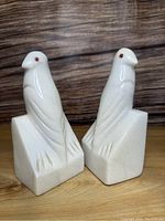 Front view of two white marble bird-shaped bookends with reddish eyes standing on a wooden surface against a wood background.