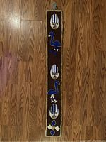 Full view of the vintage African beaded sash belt or wall hanging showing three blue bird figures, three oval medallions with faces, and a geometric flower design at the bottom, lying flat on a wood floor.