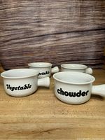 Four white ceramic soup/chowder bowls with black text labels and handles on a wooden surface.