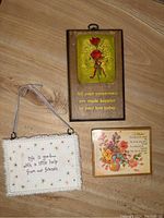 Photo of three wall decor pieces on wooden floor: a green and red flower embossed wooden plaque with gold quote, an embroidered fabric hanging with multicolor stars and quote, and a small floral card/plaque.