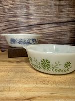 Two vintage Glasbake casserole dishes, one with a green floral pattern and the other with blue floral print, staged on a wooden surface with a wood grain backdrop.