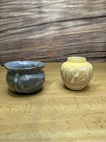 Two small pottery pots side by side on a wooden surface with a wooden textured background, showing front view.