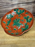 Top side of vintage Japanese ceramic plate showing red background with green horsemen, birds, and plants design and scalloped edges.