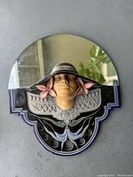 Front view of the vintage large glazed mirror showing the round mirror at the top and the sculpted woman's face with a black hat and pink flowers below it, mounted on a decorative black and blue glazed base.
