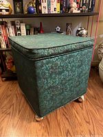 Full view of green floral patterned ottoman with padded lid, on wood floor.