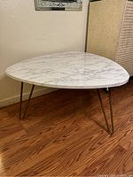 Photo of a modern triangular white faux marble coffee table with gold metal hairpin legs, shown from an angled top view on a wood floor against a beige wall.
