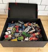 Open black box containing a collection of various vintage die-cast toy cars and trucks in assorted colors and shapes.