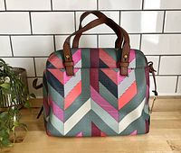 Front view of the rectangular handbag showing the multicolor chevron pattern and brown leather handles against a white tiled background.