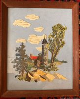 Full view of framed needlework depicting lighthouse scene with trees and rocks by the sea.