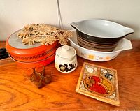 Photo of midcentury items on wood surface including Pyrex bowls, pans, amber shot glasses, pottery container, and 2 trivets