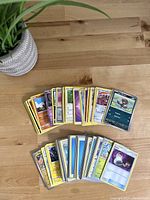 Top-down view of assorted Pokémon trading cards arranged in two rows on a wooden table with part of a plant on the left.