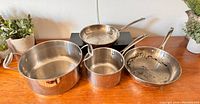 Four professional stainless steel pots and pans on wooden surface, including a large pot with handles, two saucepans with long handles, and one frying pan with wear.