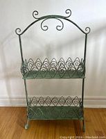 Front view of the turquoise metal magazine rack showing two shelves with diamond-pattern sides and decorative curved handle top.