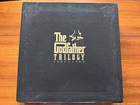 Black presentation box with gold embossed title and puppet hand logo for The Godfather Trilogy 1901-1980.