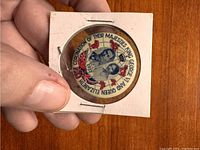 Hand holding vintage 1937 coronation button displaying King George VI and Queen Elizabeth portraits with surrounding text and British imagery.