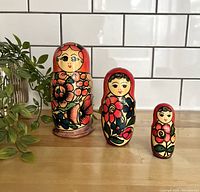 Front view of three vintage nesting dolls with red floral design on a wooden surface against a white tiled background and a small plant to the left.