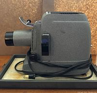 Side view of vintage slide projector showing gray textured metal body, focus knob, and cord wrapped on the base.
