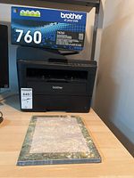 Brother MFC-L27300W printer on desk with TN760 toner cartridge box on top and a pack of paper in front