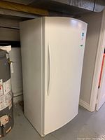 Front view of the white Danby upright refrigerator against a basement wall next to a water heater.