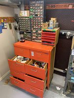 Photo showing an orange metal tool cabinet with multiple open drawers revealing various tools and materials inside, along with a portable lantern on top and two large plastic organizers with multiple small drawers containing hardware.