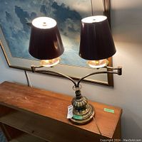 Lamp front view showing dual arms with light fixtures and black shades, decorative metal base, and approximate size on wooden surface with blue painting background.