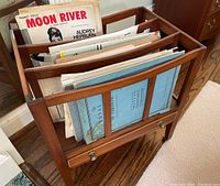 Front angle view of wood magazine rack filled with assorted sheet music including 'Moon River' piano solo for Audrey Hepburn and other vintage titles.
