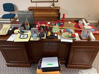 Overview of office supplies on a wooden desk including a file box, staplers, tape dispensers, flashlights, scissors, and paper products