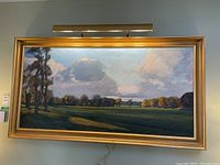 Framed oil painting with four-bulb picture light mounted above, showing full landscape scene of field, trees and sky.