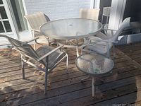 Full view of large round metal table with plexiglass top and four metal frame patio chairs with woven fabric seats and backs on a wooden deck outside near white brick wall and glass doors.