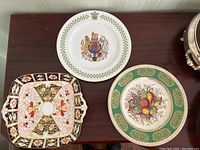 Three decorative china plates arranged on dark wood surface: one square Royal Crown Derby plate, one round Spode plate with fruit and green border, one round Ducal plate with crest and green border.