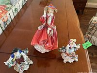 Photo showing the three figurines: one large Royal Doulton woman in red dress and two smaller child figurines with flowers seated on floral base, placed on a wooden table.