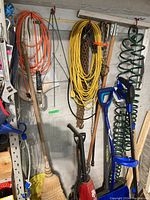 Wall display showing broom, two blue shovels, black pump, chains, cords and green coiled hose.