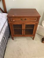 Front view of vintage style wood side table showing one drawer with metal handle and two doors with metal mesh panels on front, standing on carpet next to a mattress.