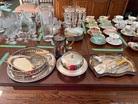 Wide shot of the silver plated trays, utensils, covered serving dish, Sheffield coffee pot, and some glassware in the background.