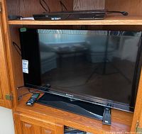 Sony 31 inch LCD TV (Model KDL-32EX340) with black bezel on wooden oak cabinet, remote on lower left corner, and Sony Blu-ray DVD player on shelf above TV.