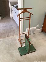 Photo showing the full wooden valet stand and green leather covered wooden leg rest on a carpeted floor next to tile flooring.